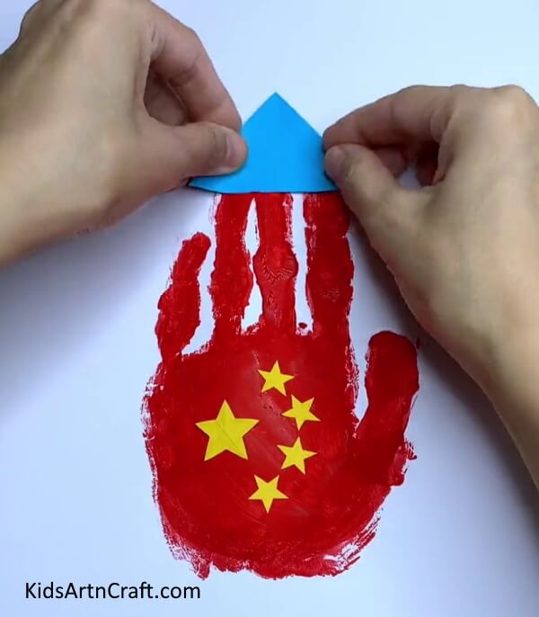 Pasting Blue Triangle On Top - Crafting a Handprint Rocket with Paper - A Step-By-Step Tutorial for Kids.