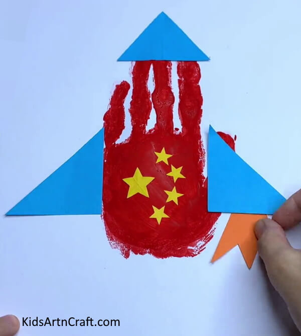 Pasting Orange Paper On the Right - Step-By-Step Guide to Making a Paper Rocket with Handprints for Kids.