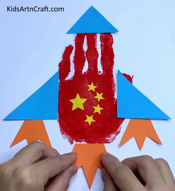 Pasting Orange Paper At the Bottom - Crafting a Paper Rocket with Handprints - A Guide for Children.