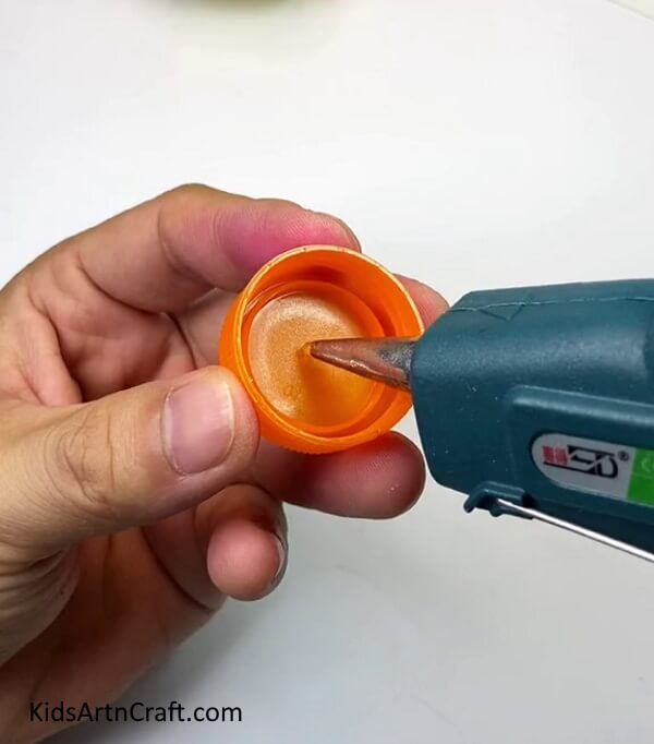 Applying Glue Gun In Caps- Crafting a toy vehicle using chopsticks, elastic bands, straws, and bottle tops.