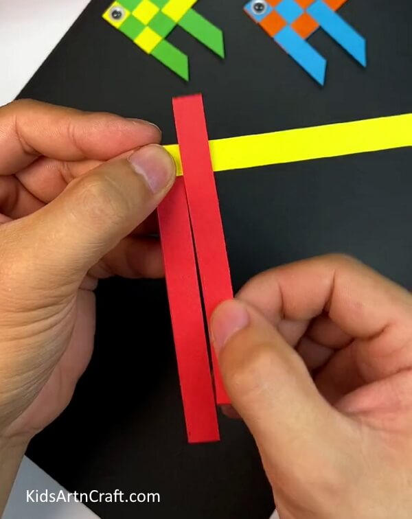 Putting the red red strip on the yellow one- Tutorial on how to make a paper fish using weaving techniques. 