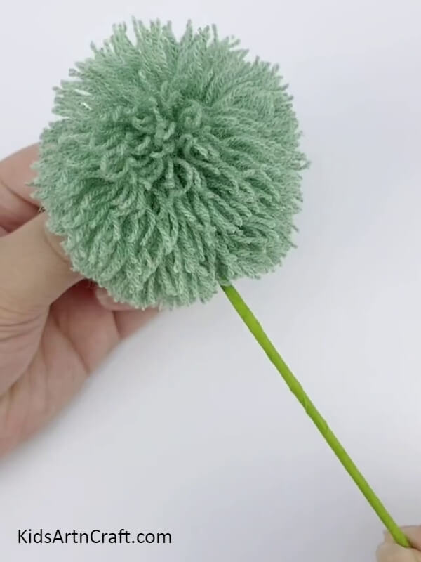 Attaching The Faux Stem To The Pom Pom Flower- Children can learn how to make Pom Pom Flowers with a variety of threads through this tutorial. 