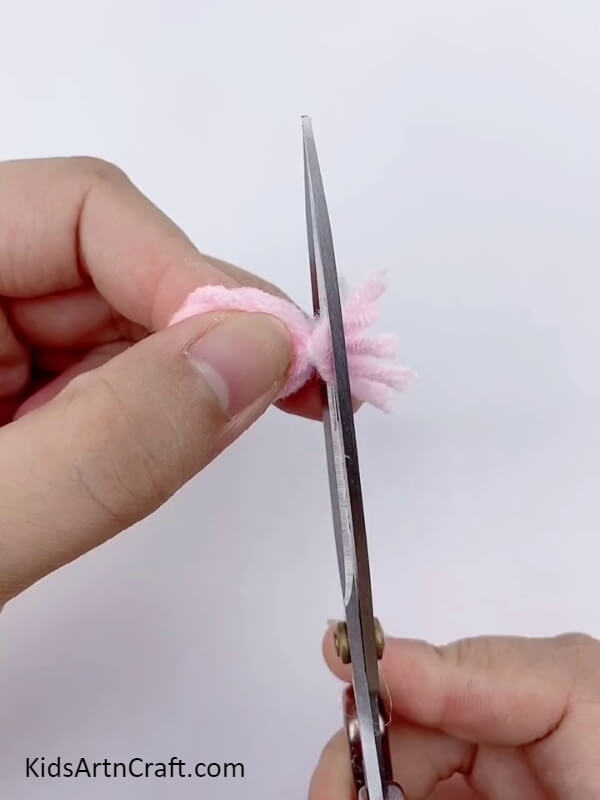 Cut the extra part of the light pink wool with scissors- Instructions for creating a beautiful floral ring with cardboard and yarn for children. 