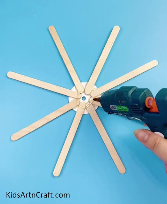 Pasting 7 More Popsicle Sticks- Introduce Children to Crafting by Utilizing Recycled Sticks and Bottle Caps to Create a Ferris Wheel. 